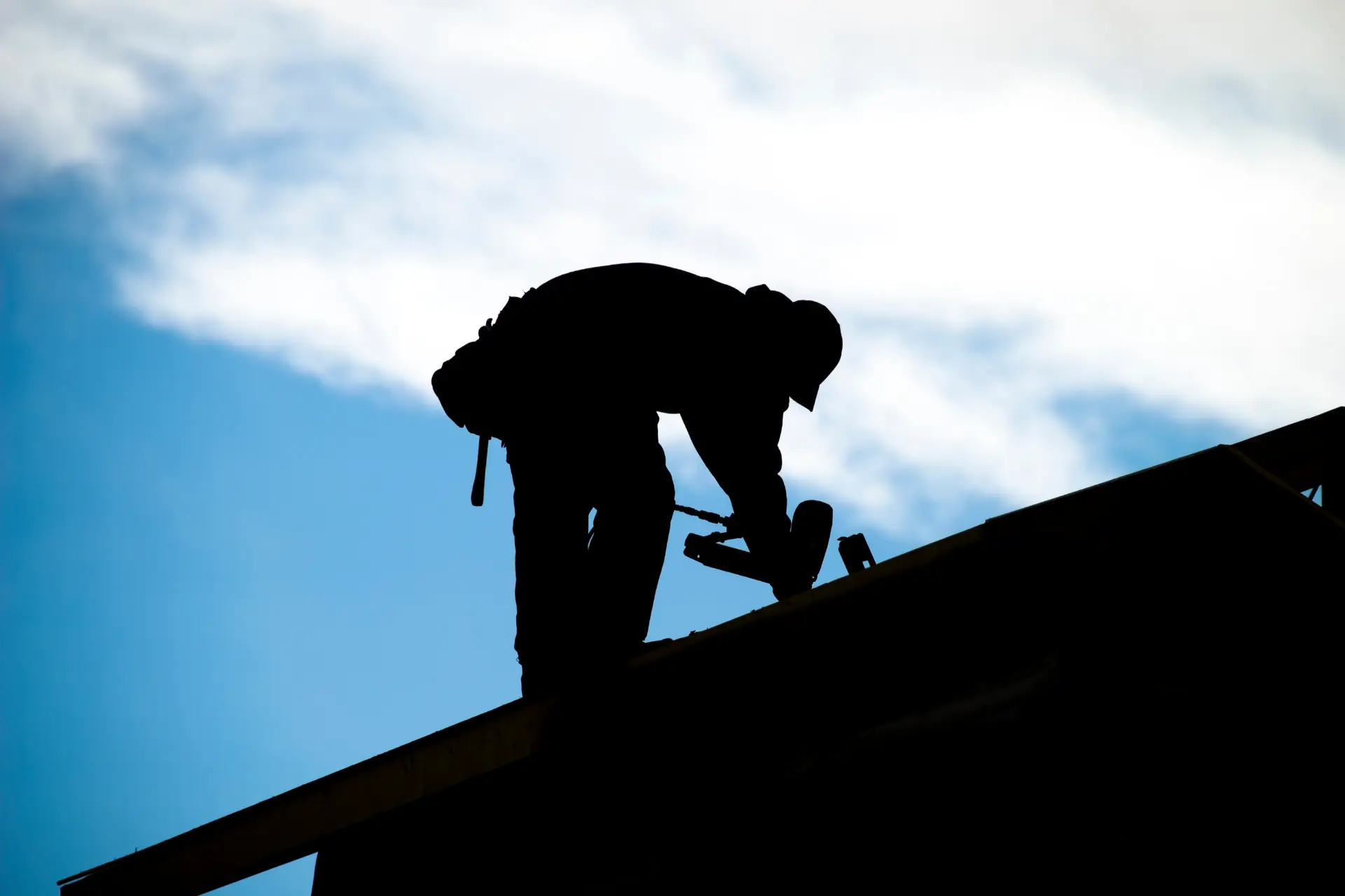 Silhouette of a roofer working on a roof ridge against a bright blue sky, representing the qualified roofing professionals homeowners should vet before signing a contract